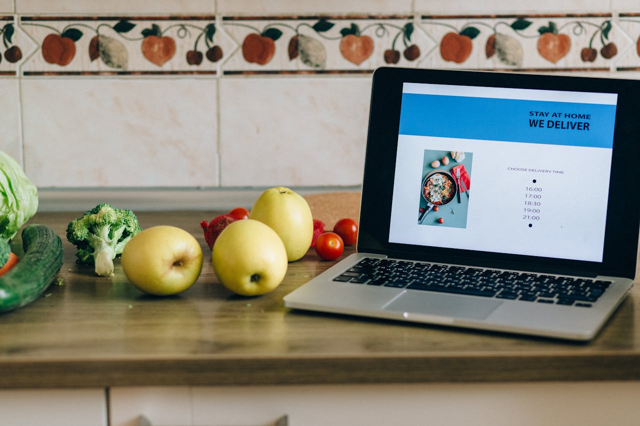 team-03 Laptop showing a delivery app beside fresh fruits and vegetables on a kitchen counter.